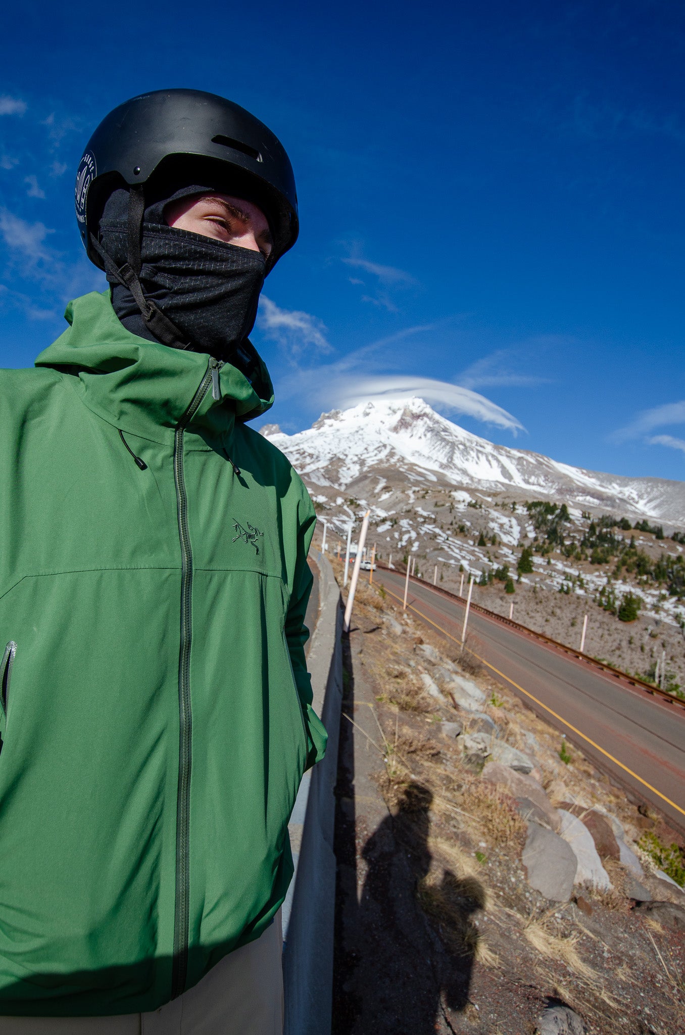 Person in green jacket and black helmet standing on a road with a snow-capped mountain in the background