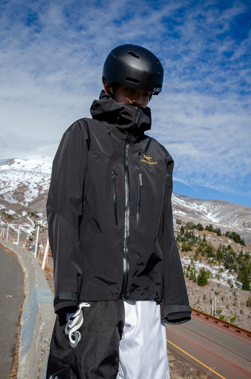 Person wearing a black jacket and helmet on a mountain road with snow-capped mountains in the background.